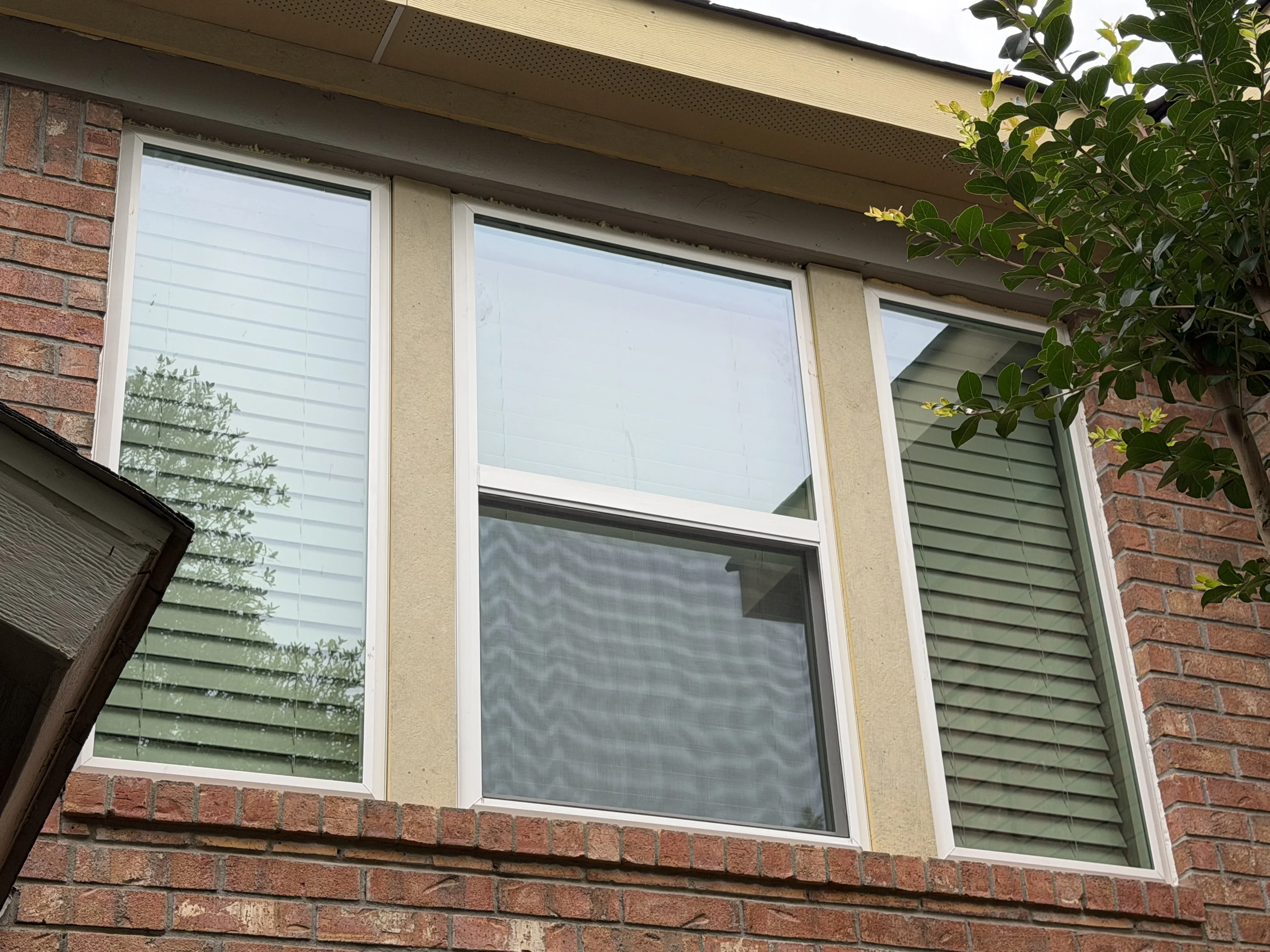 Three white-framed residential windows with horizontal blinds installed on brick home exterior